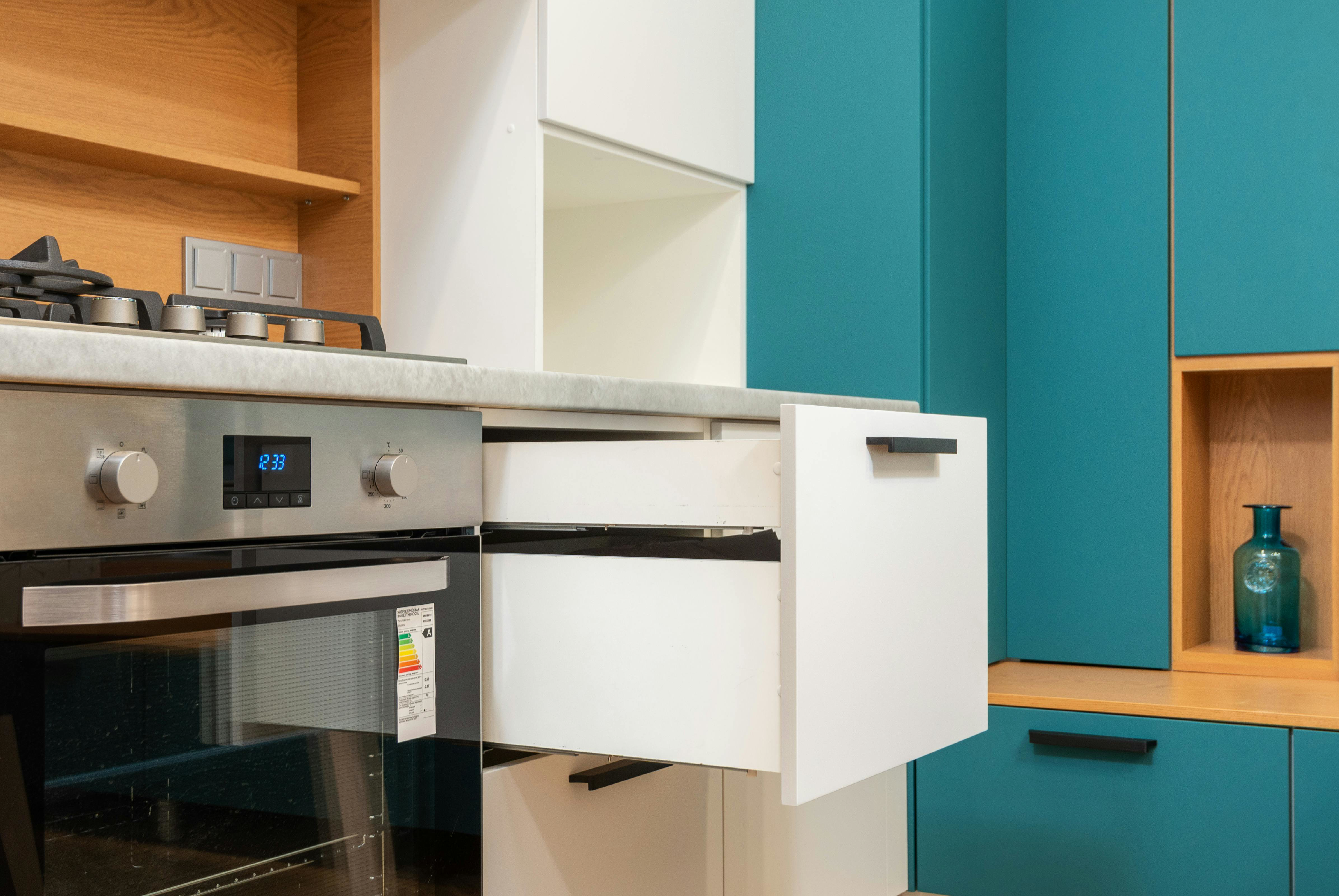 Open deep drawer beneath compact countertop in teal-and-white small kitchen, showing space-saving full-extension storage beside built-in oven.