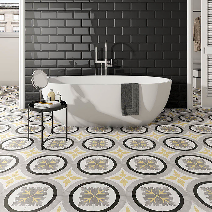  A modern bathroom featuring a white freestanding bathtub against a glossy black subway-tile wall. The floor showcases vibrant patterned tiles in circular designs with yellow and grey accents, creating a bold contrast with the dark backdrop. A small black side table with a mirror and bath essentials adds a stylish touch.