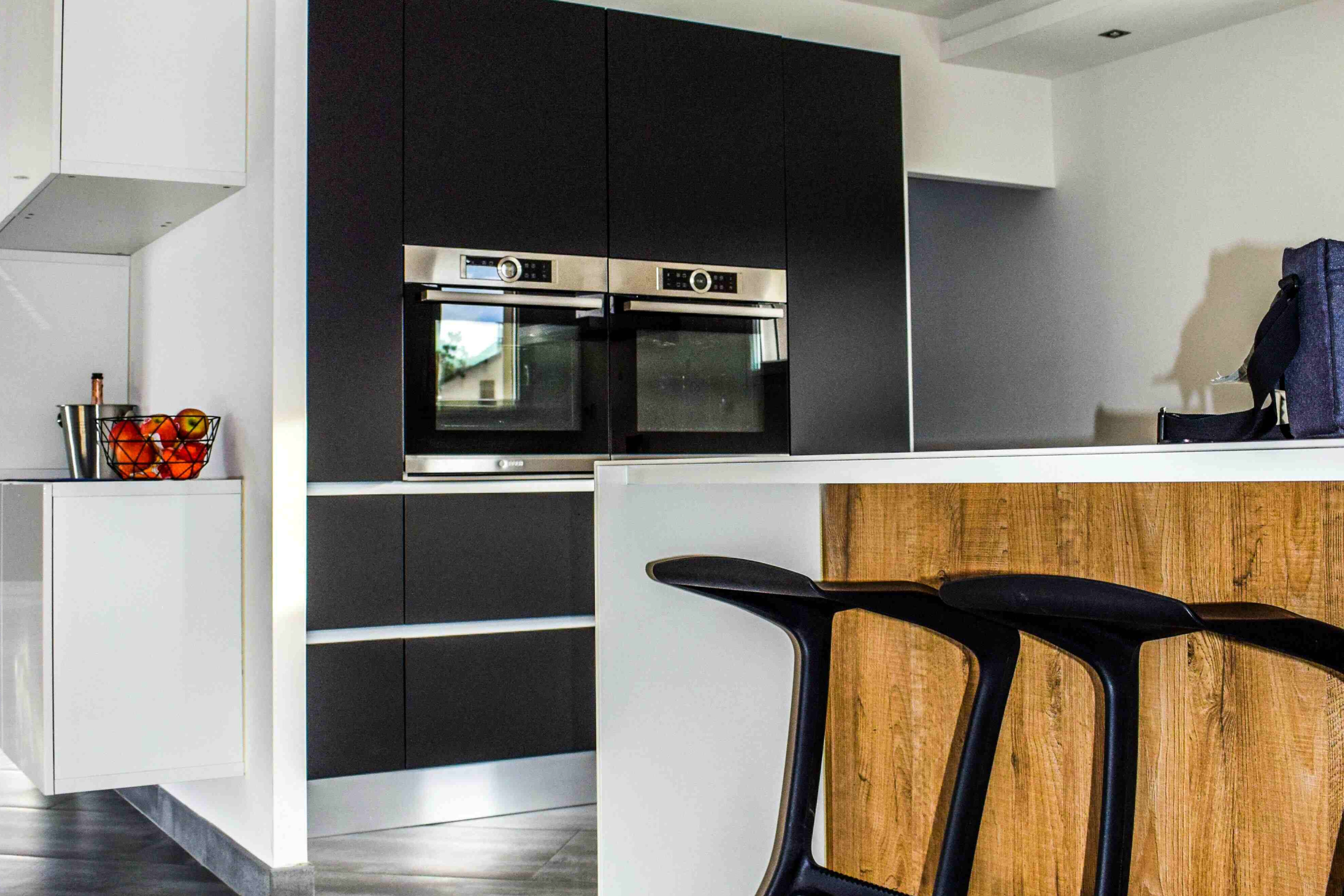Small modern kitchen with tall matte-black cabinets, double built-in ovens, white island with wood front, slim black bar stools and grey floor.