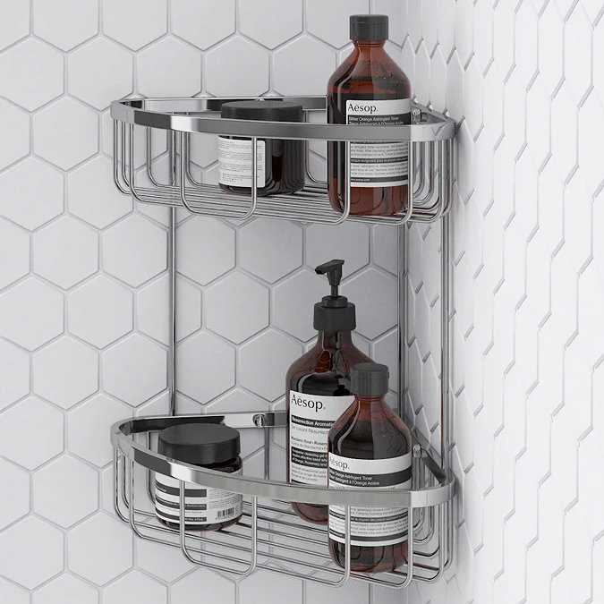 Chrome Toreno corner shower caddy with two wire-frame shelves, holding amber Aesop bottles and tubs, mounted on hexagonal white tiles.