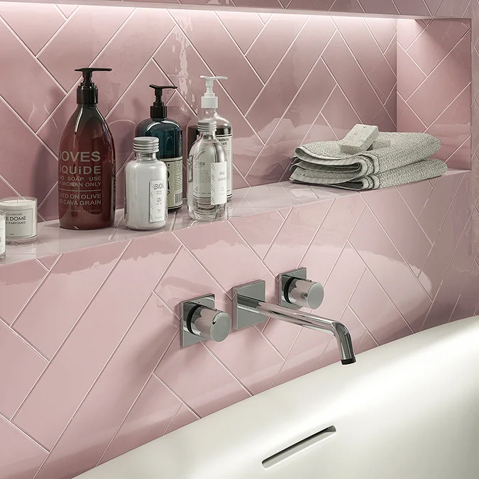 Close-up of a stylish bathroom with pink herringbone tiles, a recessed shelf with toiletries, and a chrome wall-mounted tap over a white bathtub.