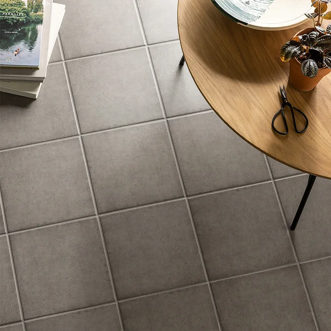 Matte grey square floor tiles paired with a wooden round table and potted plant, creating a cosy and modern interior space.