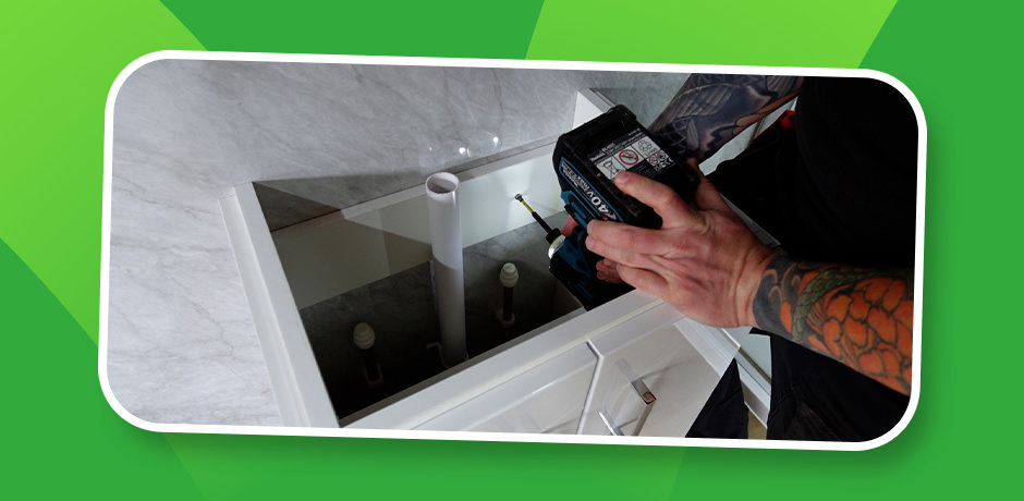 Installer driving a screw through the rear rail to secure a white floor-standing vanity unit to a marble-effect bathroom wall, with hot, cold and waste pipes already fed through the cabinet