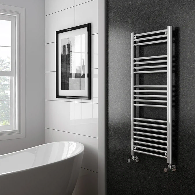 Modern bathroom featuring a chrome heated towel rail mounted on a dark wall next to a freestanding bath, with large white tiles and framed wall art near a bright window.