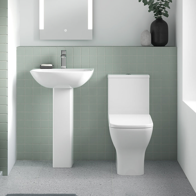 Minimalist bathroom with white pedestal sink, modern toilet, LED mirror, and soft green tiled backsplash.