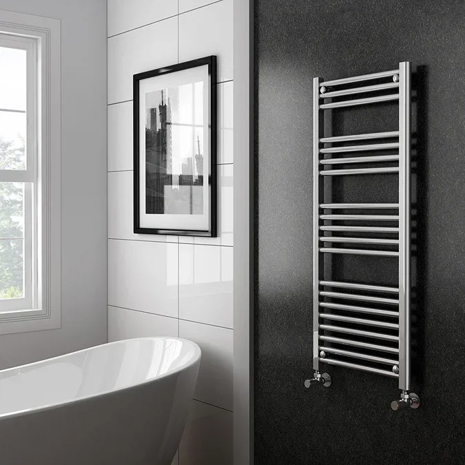 Modern bathroom featuring a chrome heated towel rail mounted on a dark wall next to a freestanding bath, with large white tiles and framed wall art near a bright window.