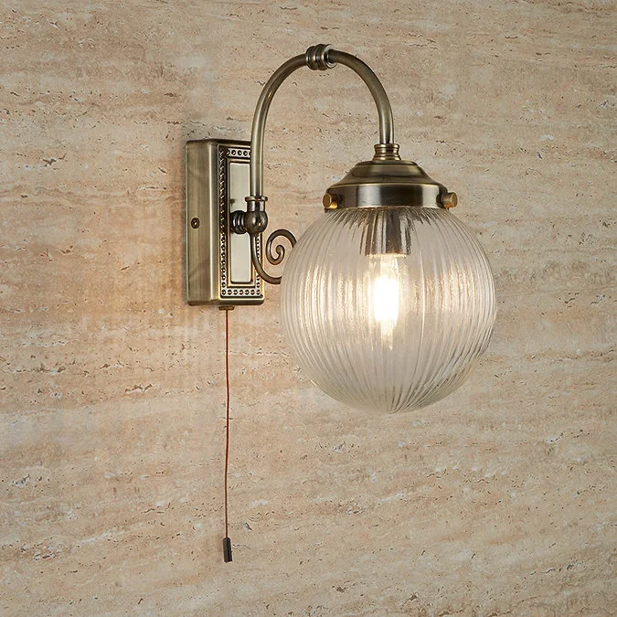 Antique brass wall light with a ribbed glass globe shade and decorative curved arm, mounted on a beige textured wall, featuring a pull switch for easy operation.