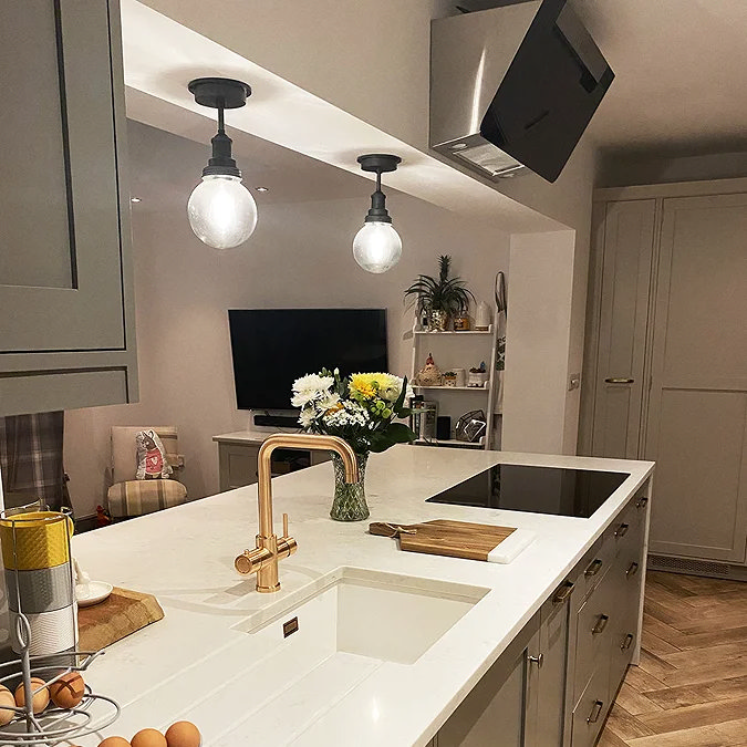 Modern kitchen with industrial-style pendant lights featuring clear glass globes, a white quartz island countertop with a gold mixer tap, and an open-plan living space in the background.