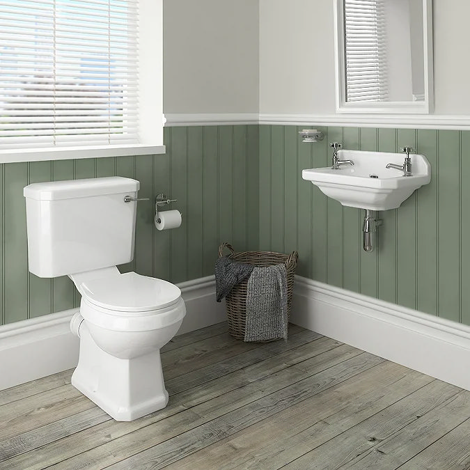 Traditional bathroom with sage green wood panelling, featuring a classic white toilet and a corner wall-mounted basin. The room includes a woven laundry basket with a towel and light wood flooring, creating a rustic, serene atmosphere.