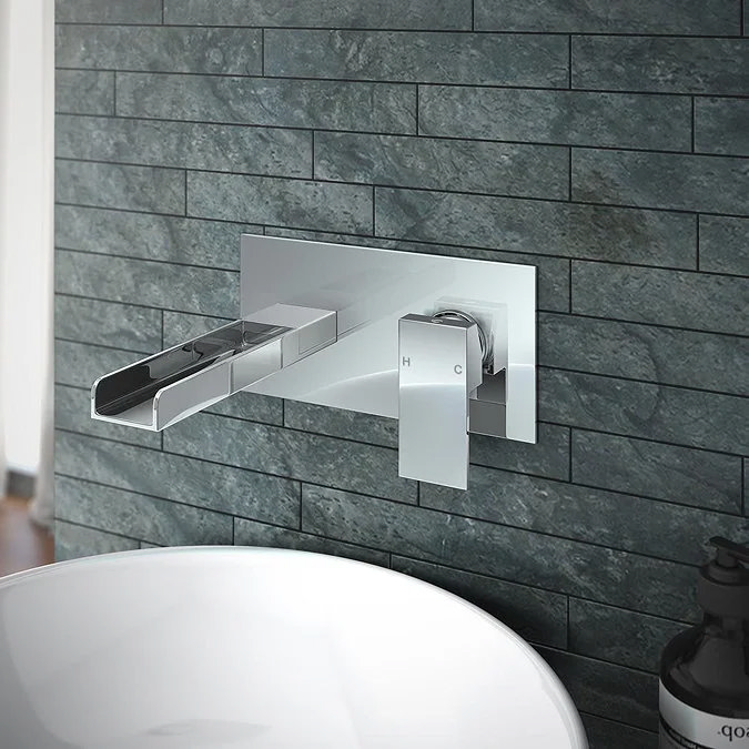 Modern wall-mounted chrome waterfall tap with sleek handle set against textured dark tiles, adding a touch of luxury above a minimalist white basin.