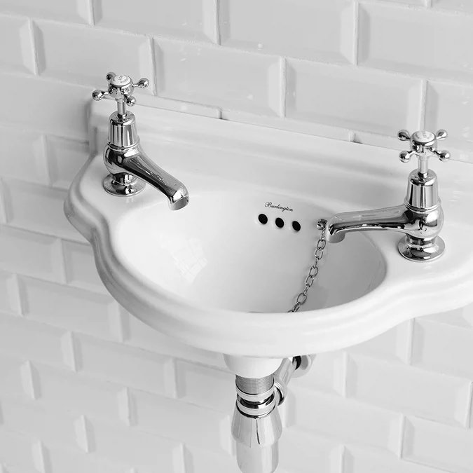 Small traditional wall-mounted white cloakroom basin with two chrome pillar taps and ceramic cross handles against white tile wall.