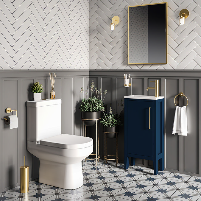 Stylish bathroom with white herringbone wall tiles and grey wainscoting, featuring a white toilet, blue vanity unit with gold faucet, and decorative plants in black and gold planters. The floor displays a geometric patterned tile, complemented by gold accents on fixtures and a round wall mirror.