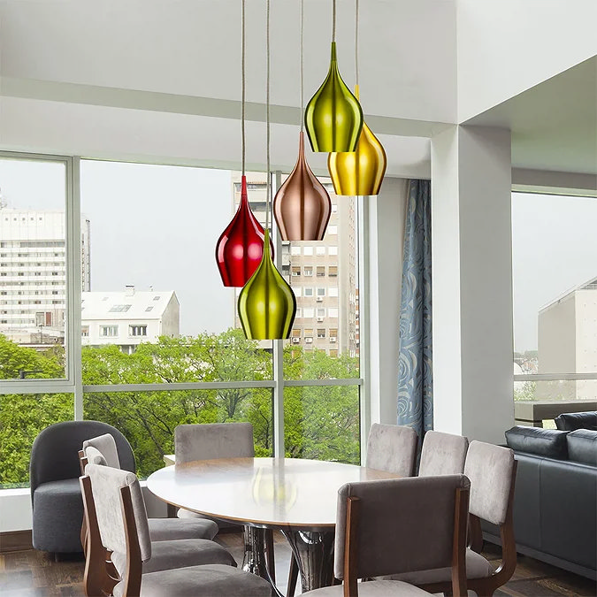 Modern dining area with a cluster of colourful, teardrop-shaped pendant lights in green, red, and gold hues, hanging above a round wooden dining table with grey chairs, set against a large window with an urban view.