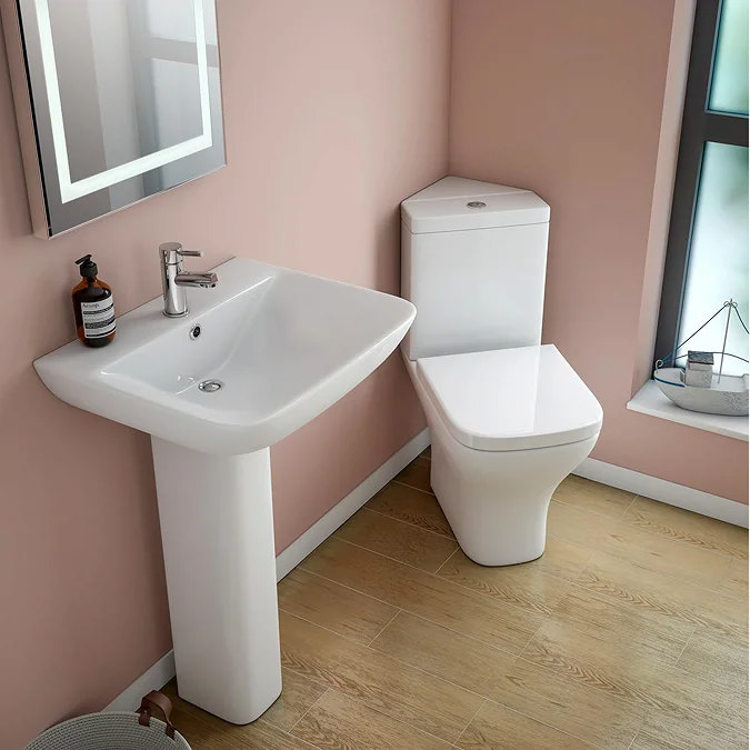 Space-saving corner toilet and pedestal basin in a small bathroom with light pink walls and wood-effect flooring