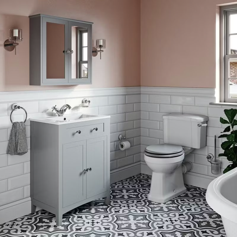 Charming bathroom with soft pink walls, grey vanity unit, and a classic toilet. Features white subway tiles, a patterned black and white floor, and a mirrored cabinet for a modern vintage look.