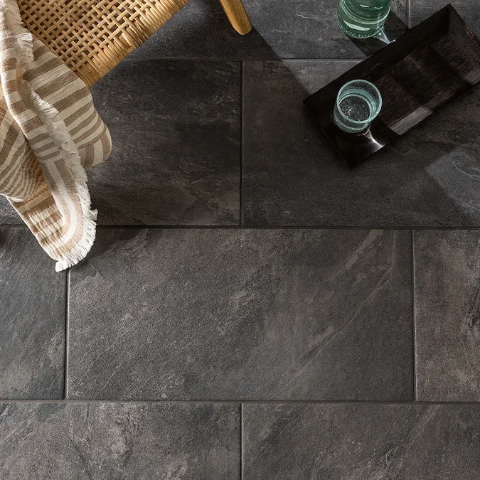 Dark stone-effect floor tiles with a textured finish, paired with a wicker chair, striped towel, and tray with a glass, creating a cosy and natural look.