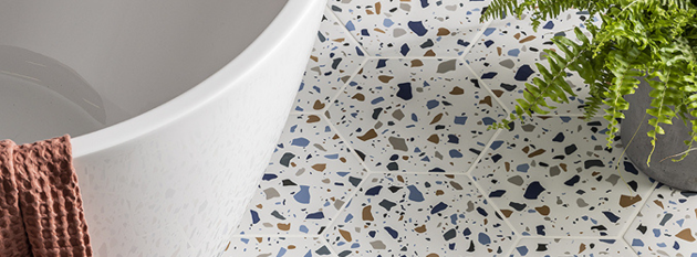 Terrazzo-style bathroom floor tiles with white background and scattered blue, brown, and black chips beside a modern white freestanding bath