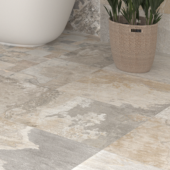 Close-up of a rustic bathroom floor featuring stone-effect tiles in soft, neutral tones. The textured tiles showcase a mix of beige, grey, and earthy hues, adding a natural, rugged charm to the space. A potted plant and the base of a freestanding bathtub complement the warm, organic feel.