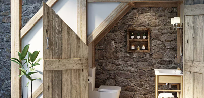 Rustic country-style bathroom with exposed stone walls, natural wood doors, and wooden beams. Features a simple white toilet, sink with wooden vanity, and greenery for a cosy, earthy feel.