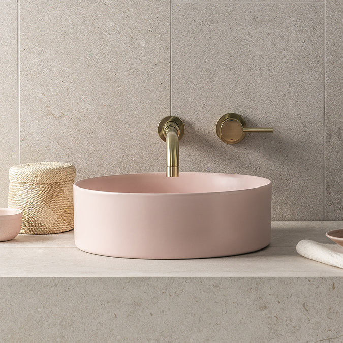 Modern bathroom with a blush pink freestanding vessel sink, brushed brass wall-mounted taps, and a natural stone backdrop for a muted, spa-like aesthetic.