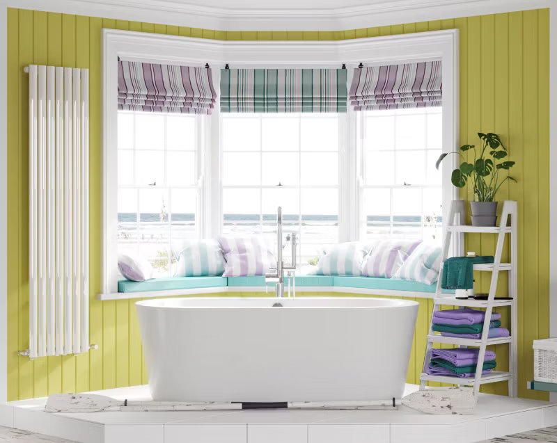 Bright and airy bathroom featuring a freestanding bathtub in front of a large bay window with coastal views, pastel striped cushions, and a cheerful yellow panelled wall for a relaxing seaside vibe.