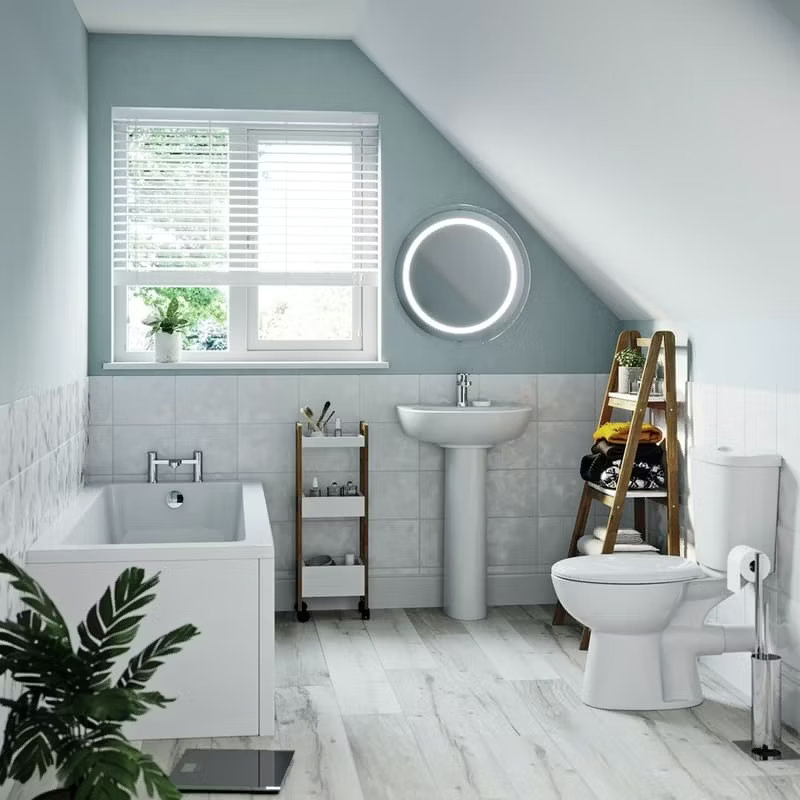 Bright and airy bathroom with sloped ceiling, featuring a pedestal sink, modern toilet, and a bathtub, showcasing efficient use of space under a window with light wood flooring and soft blue walls.