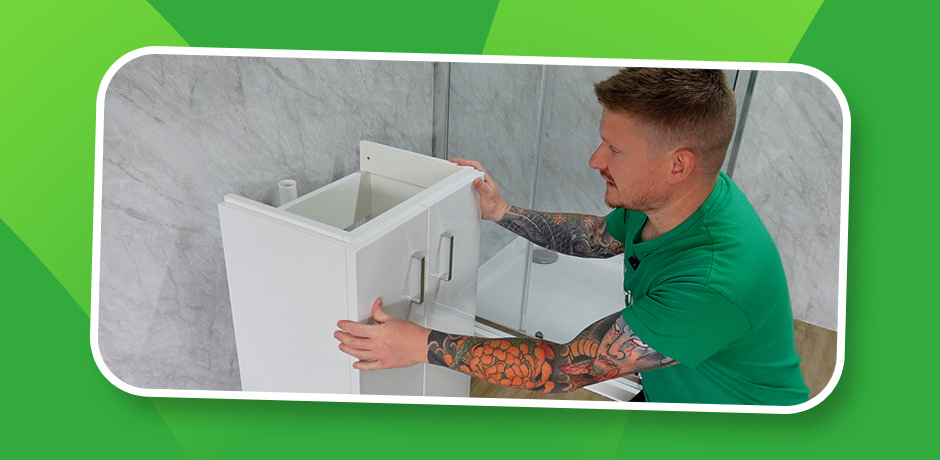 Installer slides assembled white vanity unit with doors into final position against tiled bathroom wall after drilling pipe cut-outs