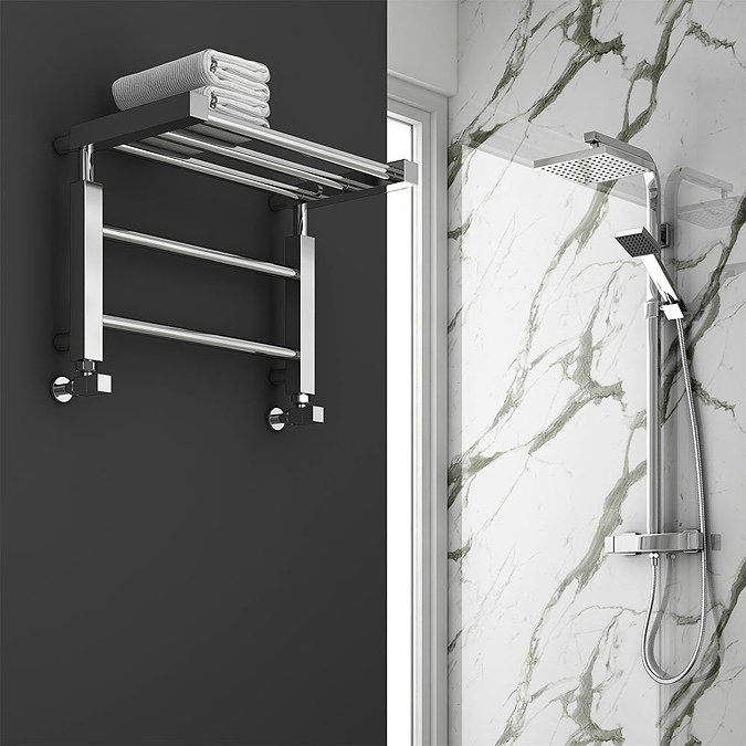Chrome heated towel rail with top shelf holding folded towels beside a marble shower wall