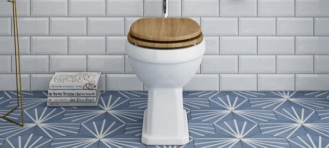 Classic white toilet with wooden seat, placed on a patterned blue tiled floor with white subway tile backdrop, illustrating ideal UK toilet dimensions for comfort and space efficiency.