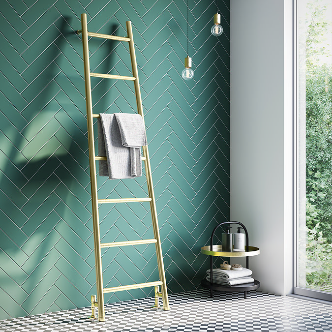 Contemporary bathroom with a gold towel ladder against a green herringbone tile wall, featuring black and white checkered flooring and a side table with bathroom essentials near a large window.