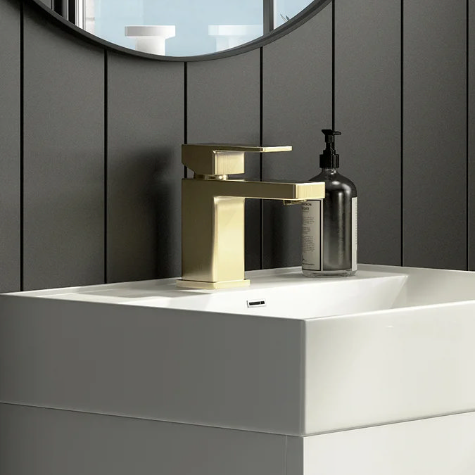 Modern brushed brass Toreno basin mixer tap on a white square basin, set against dark vertical panelled wall with circular mirror and black soap dispenser