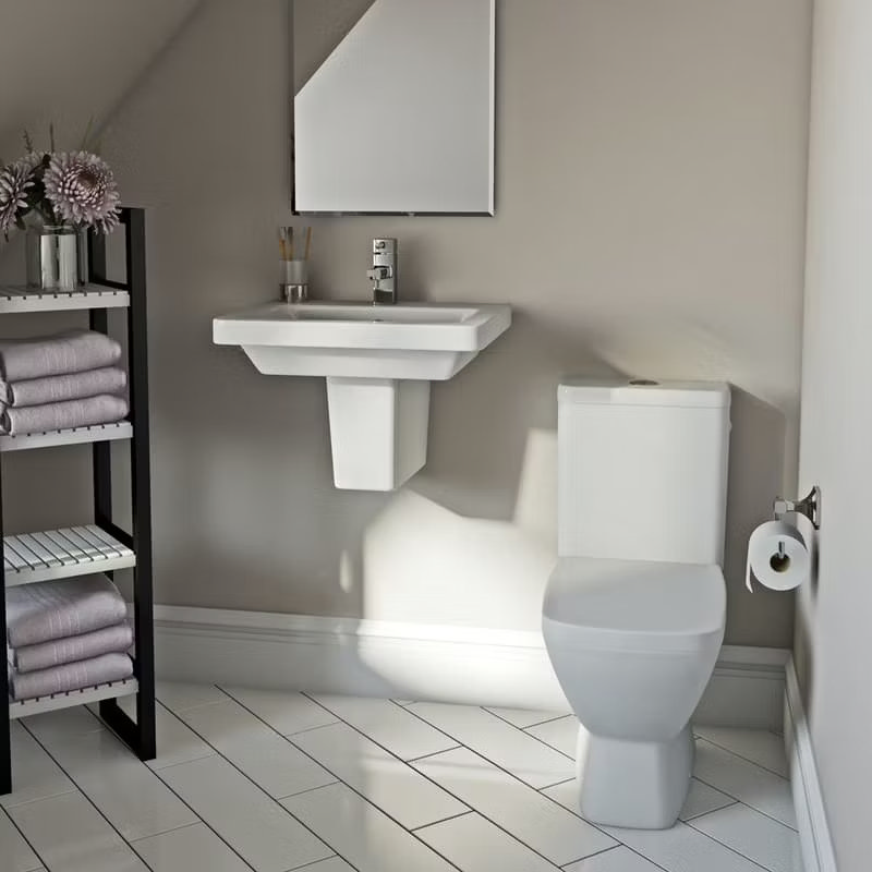 Minimalist bathroom featuring a sleek pedestal sink and modern toilet. Bright neutral tones, herringbone floor tiles, and open shelving with soft towels complete the stylish look.