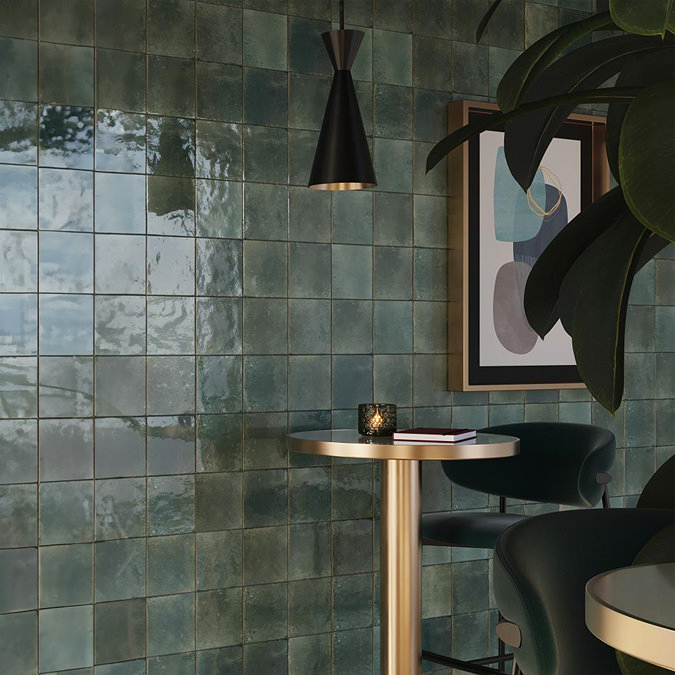 Glossy green square wall tiles with a handcrafted, rustic texture create an elegant, reflective surface in a modern interior, featuring a black pendant light, gold-trimmed table, and abstract wall art.