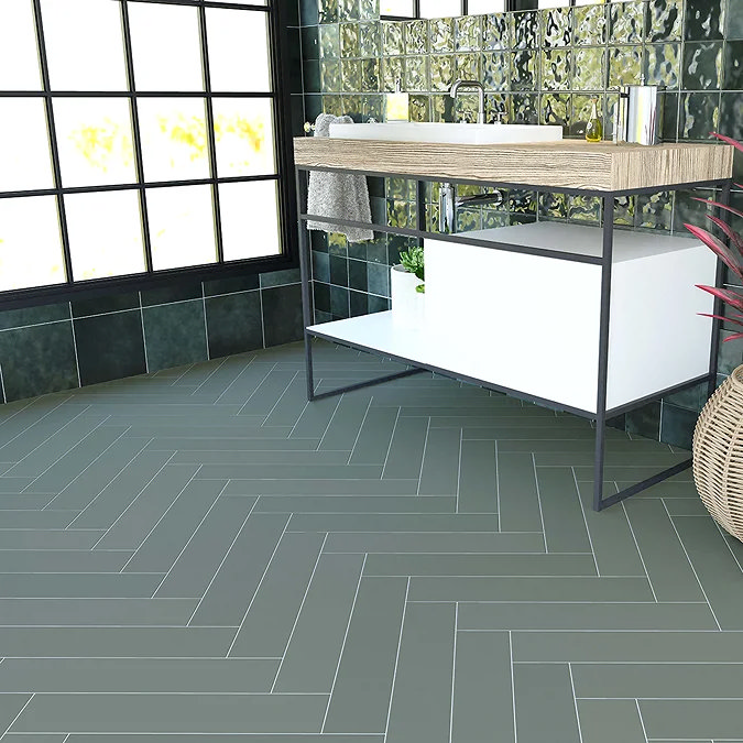 Green herringbone pattern floor tiles in a modern bathroom with a wood and metal vanity, glass panel walls, and lush indoor plants.