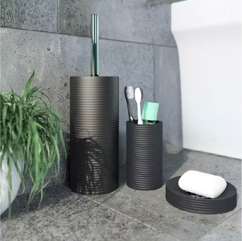 Modern black bathroom accessories set including toilet brush, toothbrush holder, and soap dish on a grey tiled floor, with a touch of greenery.