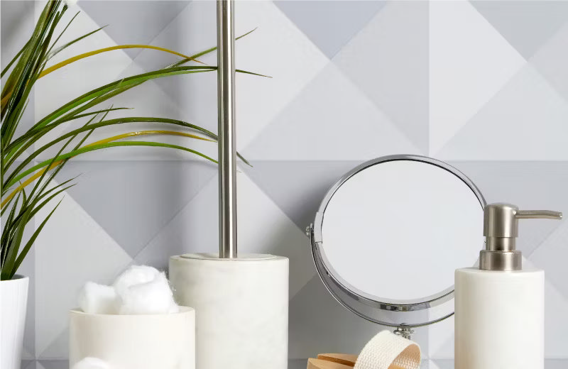 Modern bathroom accessories with a marble toothbrush holder, soap dispenser, cotton jar, and a chrome mirror, complemented by a green plant against geometric patterned tiles.