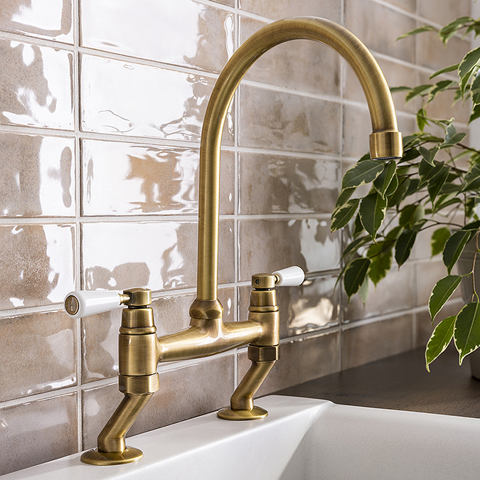 Brushed brass vintage-style tap with white ceramic handles, mounted on a glossy beige textured tile backsplash, complemented by lush green foliage.