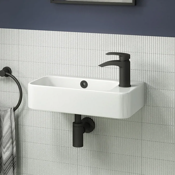 Rectangular white wall-hung cloakroom basin with black mono tap against light textured tiles.