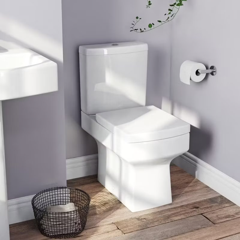 Modern close-coupled toilet with square edges in a minimalist bathroom featuring light purple walls, wooden flooring, and a wire basket for towels.