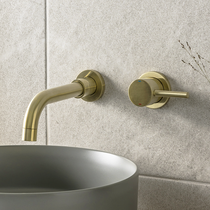 Close-up of a modern brass wall-mounted tap and mixer above a matte grey vessel sink, set against a neutral stone wall in a minimalist bathroom.