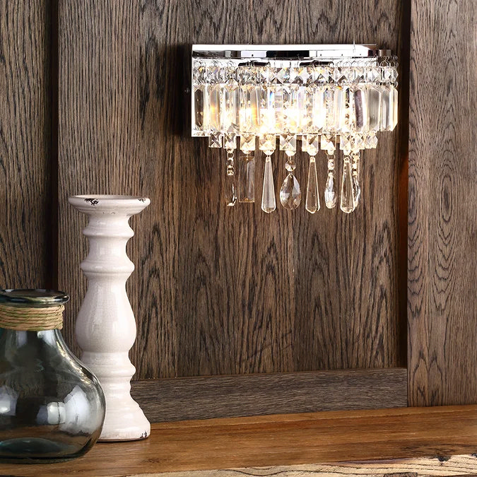 Crystal wall light with cascading glass droplets and a polished chrome base, mounted on a rustic wood-paneled wall, styled with decorative vases and a white candlestick.