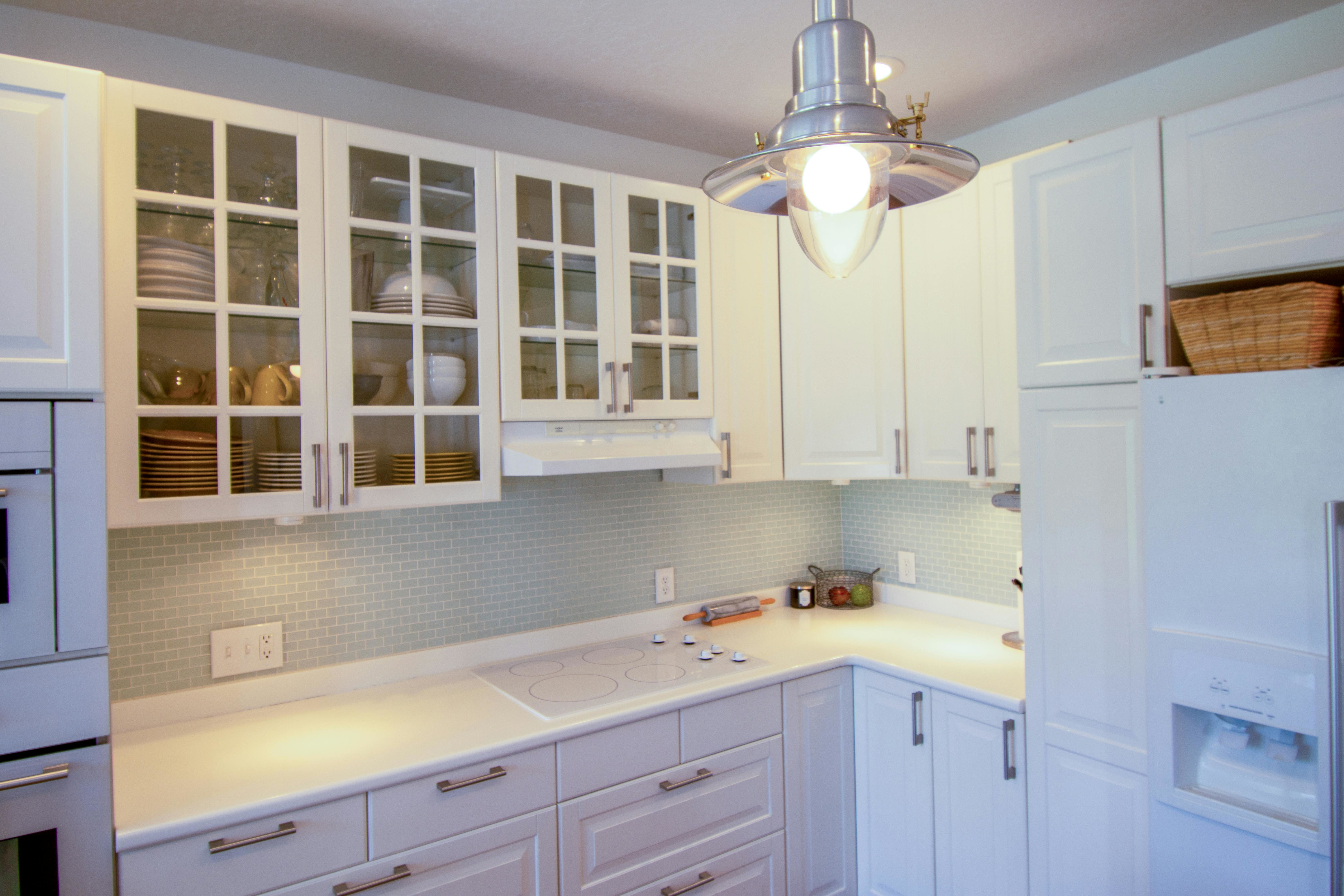 Small L-shaped kitchen with white glass-front cabinets, pale blue mosaic splashback, pendant and under-cabinet lighting—light, reflective decor for a small kitchen.