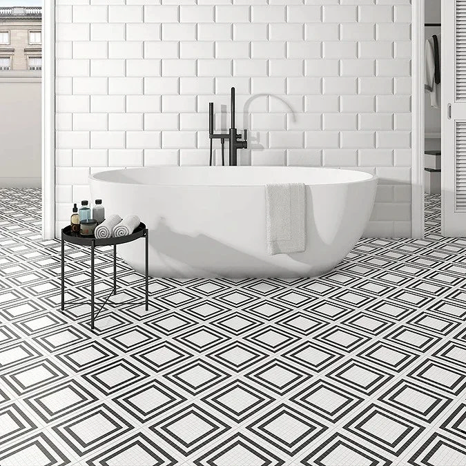 Freestanding white bathtub with black fixtures, set against white subway tiles and bold black-and-white geometric patterned floor tiles in a modern bathroom.