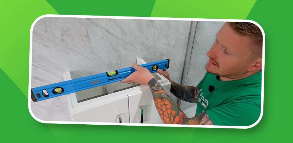 Bathroom fitter checking Step 3 of vanity installation, holding blue spirit level on top of white unit to ensure cabinet sits perfectly level against tiled wall