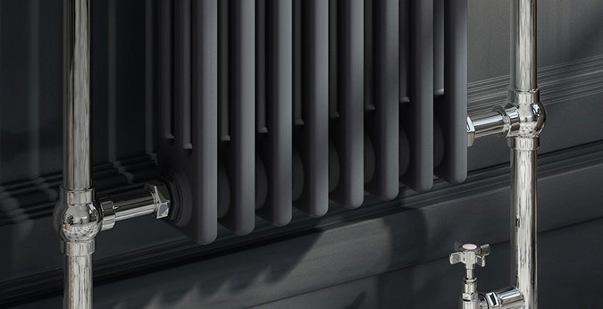 Close-up of a black column radiator with polished chrome angled radiator valves fitted to floor pipework.