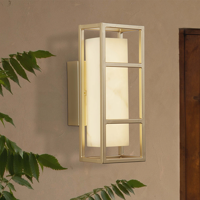 A matt gold rectangular wall light with a glowing marble‑effect diffuser mounted on a textured wall beside a wooden door and leafy plant.