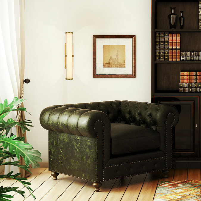 A tall gold wall light with a glowing ribbed opal glass diffuser mounted on a light wall beside a dark green armchair, a framed artwork, and a bookcase.