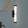A matt black clamp‑style wall light with a softly glowing ribbed opal glass tube mounted on a grey bathroom wall beside a round mirror and black tap.