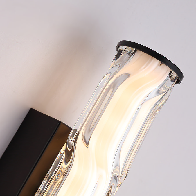 Close‑up of the wall light showing the curved wave‑ridged glass diffuser, matt black backplate, and the matt black end cap illuminated from within.