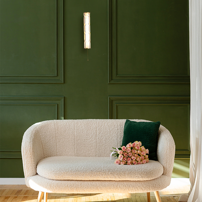 A tall matt gold wall light with a glowing wave‑ridged glass diffuser mounted on a dark green panelled wall above a cream upholstered sofa with a green cushion and a bouquet of flowers.